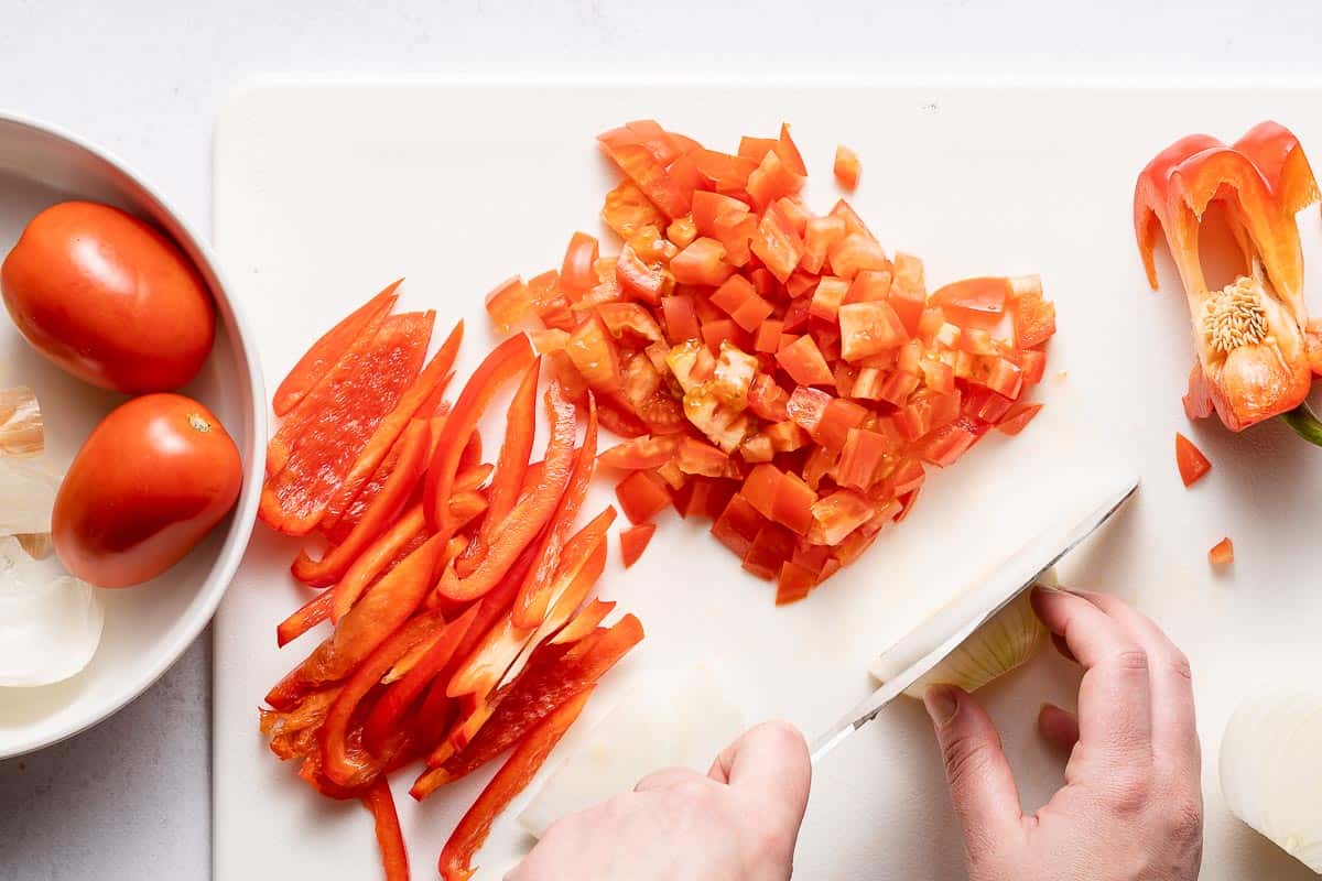 onion being chopped on cutting board with bell pepper and tomato on the side