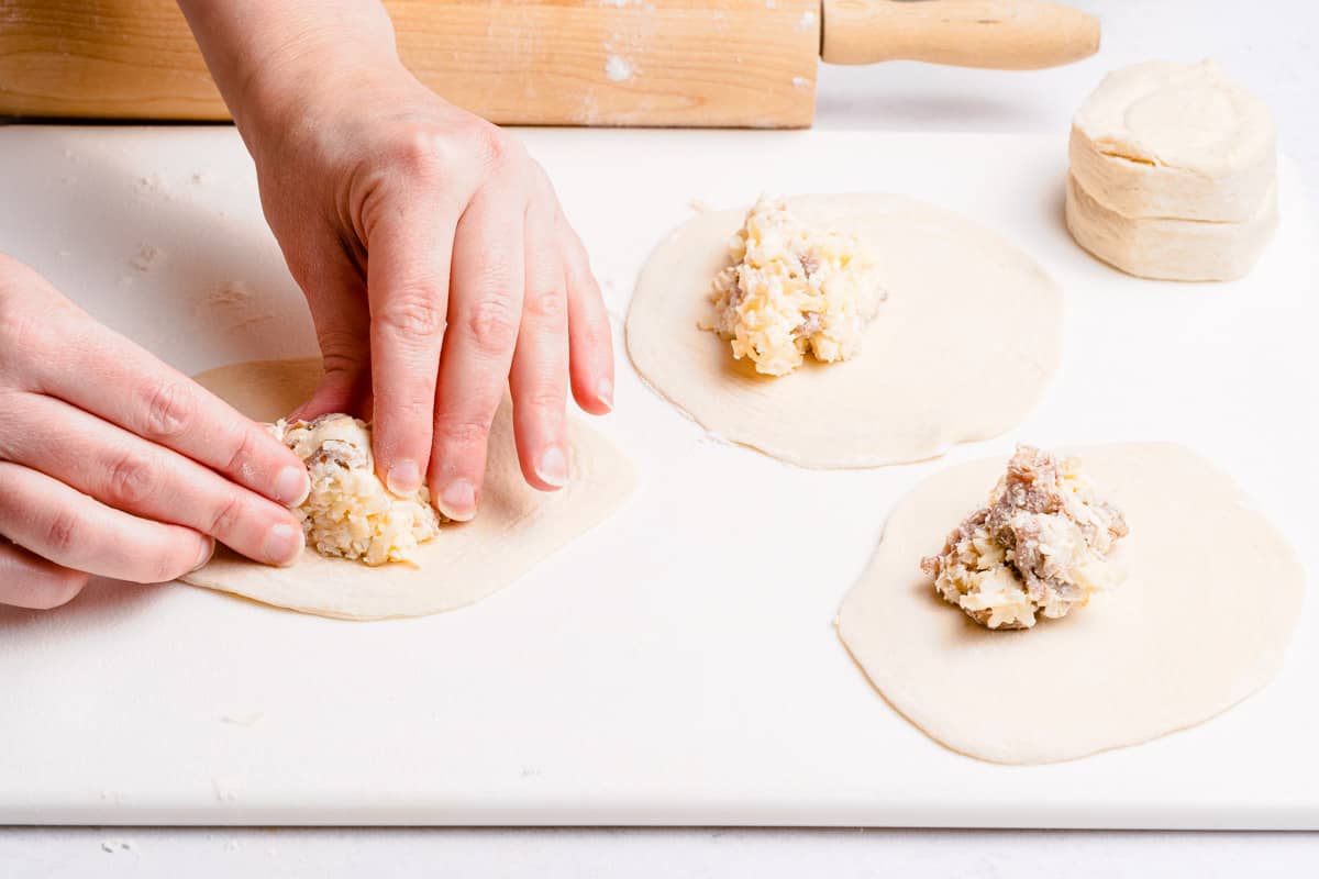 hands pressing mound of sausage and cheese filling to center of biscuit dough