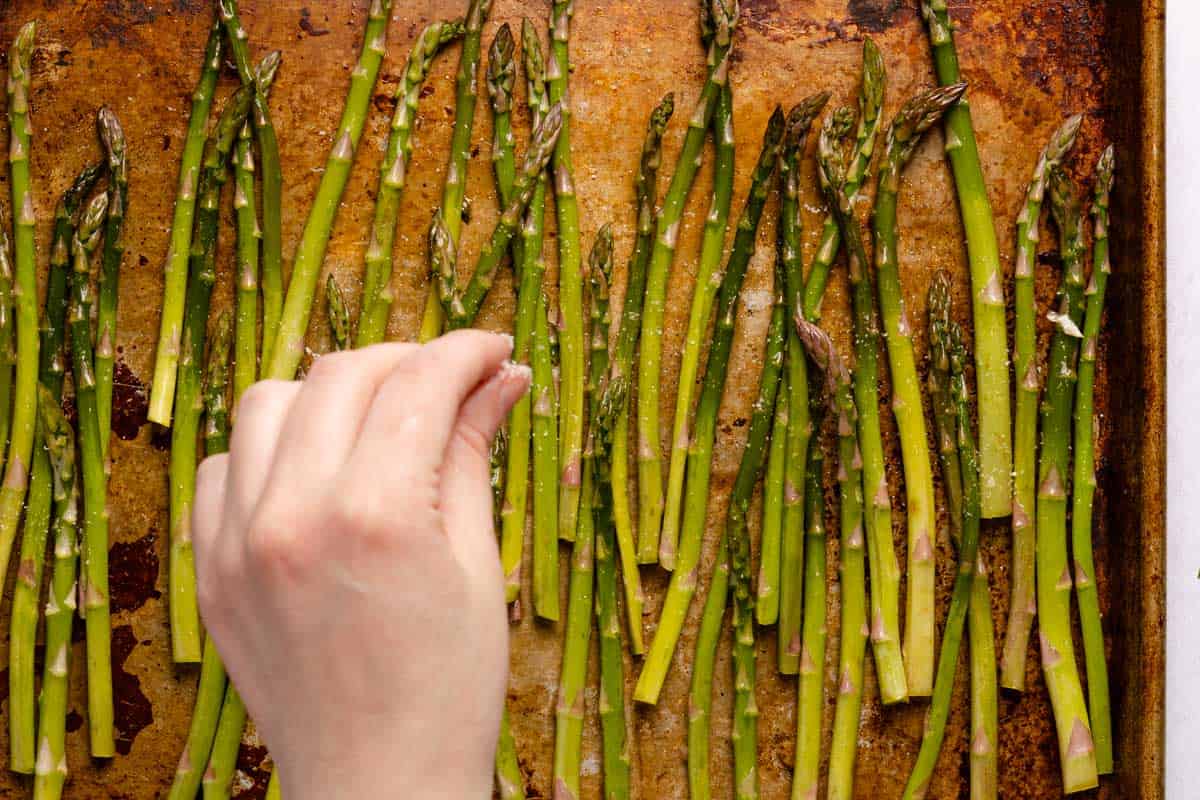 hand sprinkling salt on asparagus laid out on baking pan