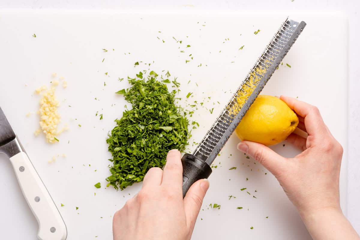lemon being zested with rasp on cutting board with herbs and garlic on the side
