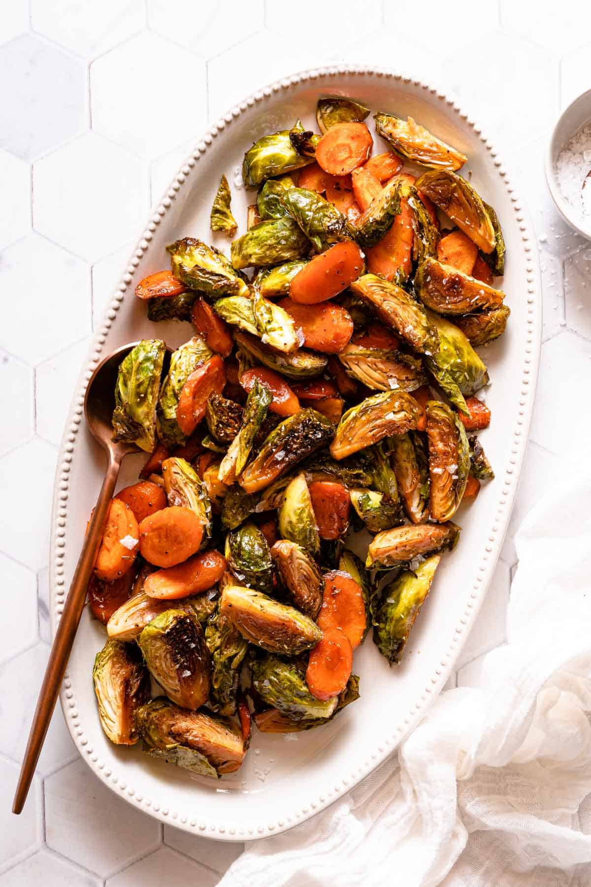roasted carrots and  brussels sprouts on white platter with spoon on the side