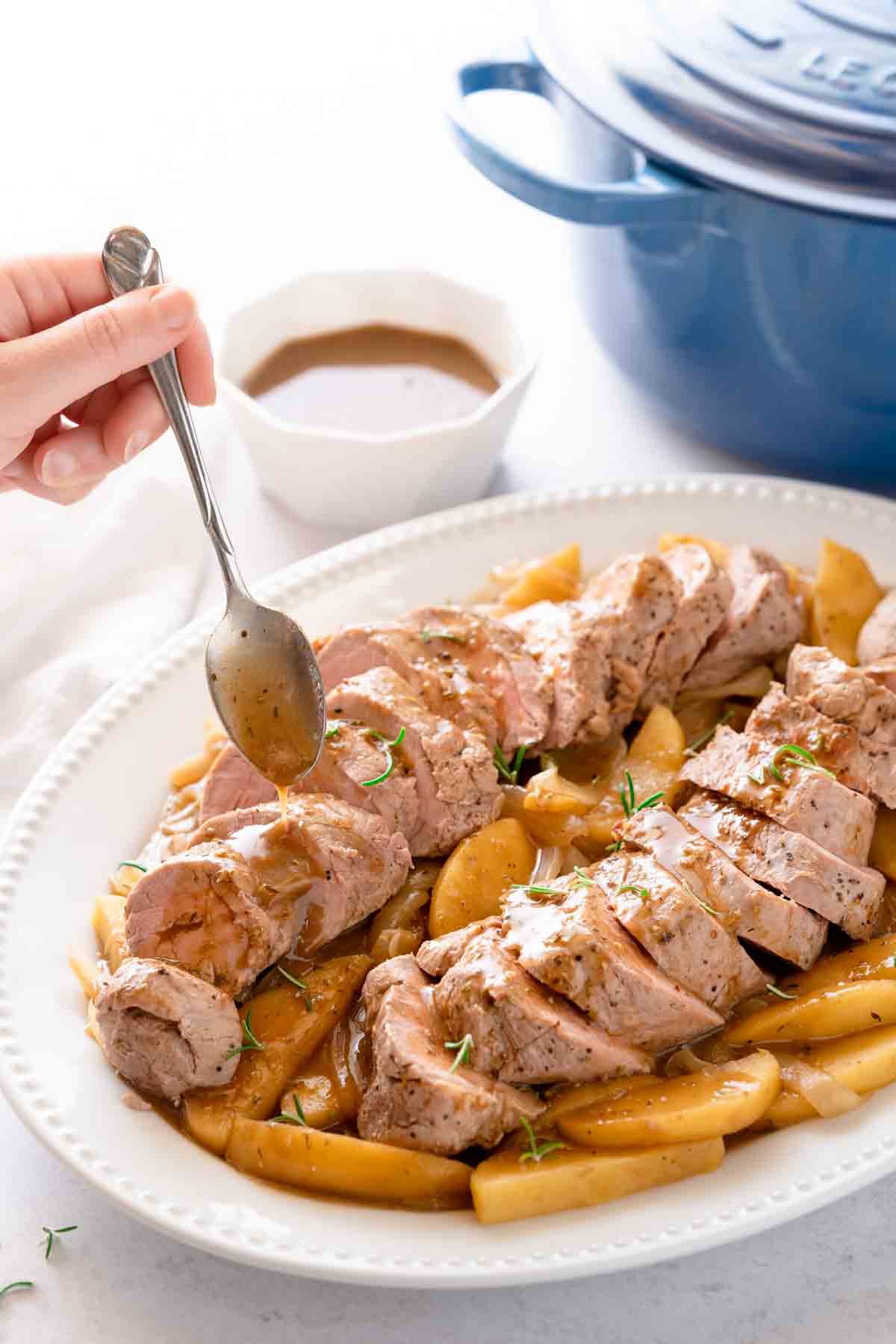 platter of sliced pork tenderloin, apples and onions being drizzled with gravy from spoon, with a Dutch oven in the background.