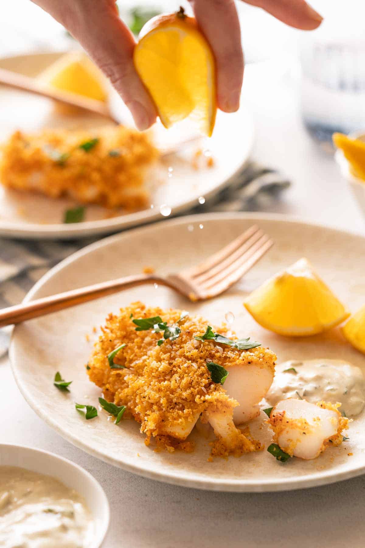 lemon being squeezed over panko crusted cod with tartar sauce and lemon on the side.