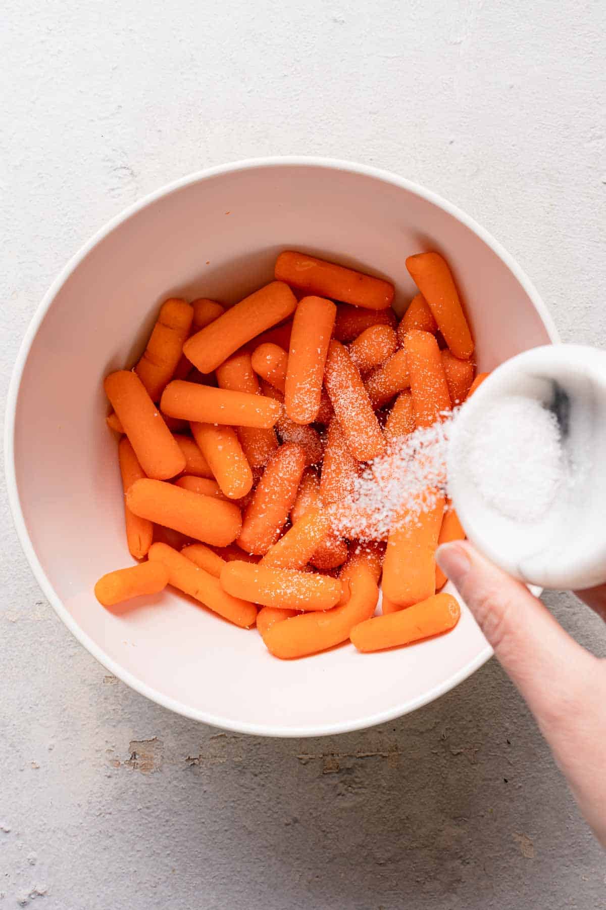 salt being sprinkled from small dish onto baby carrots in a bowl.