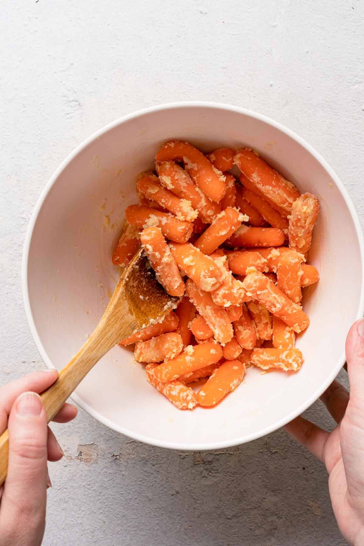 wooden spoon mixing butter with baby carrots in a bowl.