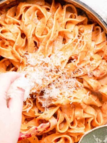 hand sprinkling Parmesan cheese over pan of pink pasta.