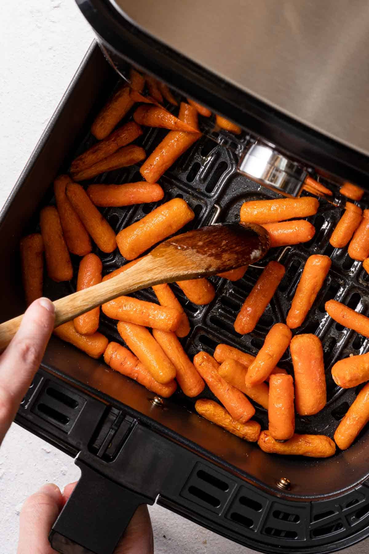 half cooked baby carrots being stirred in air fryer with wooden spoon.