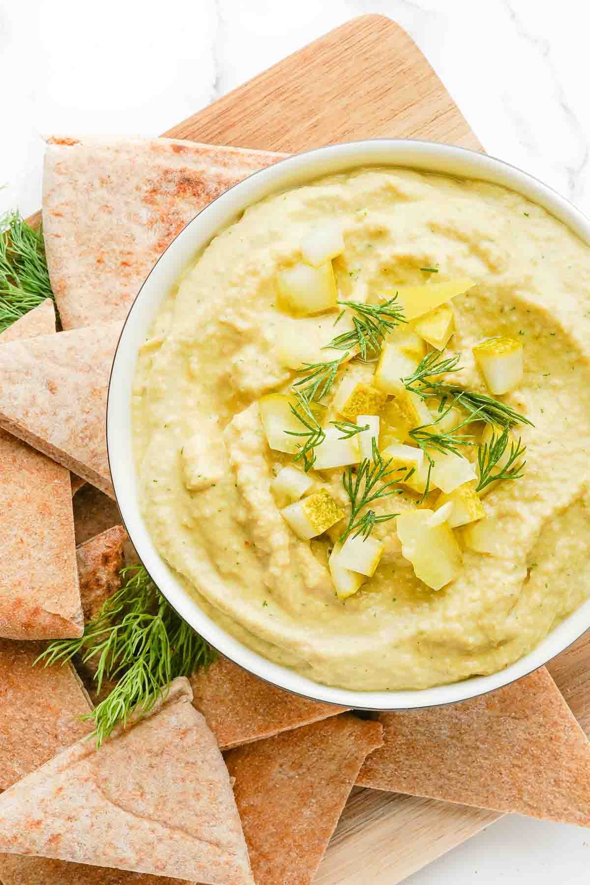 bowl of hummus garnished with pickles and dill with pita on the side.