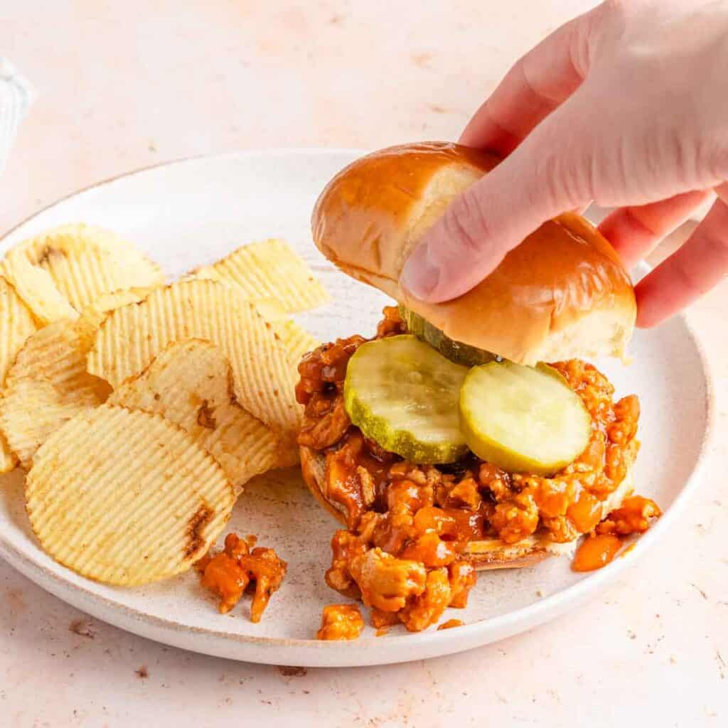 hand adding bun top to sloppy Joe sandwich with chips on the side.