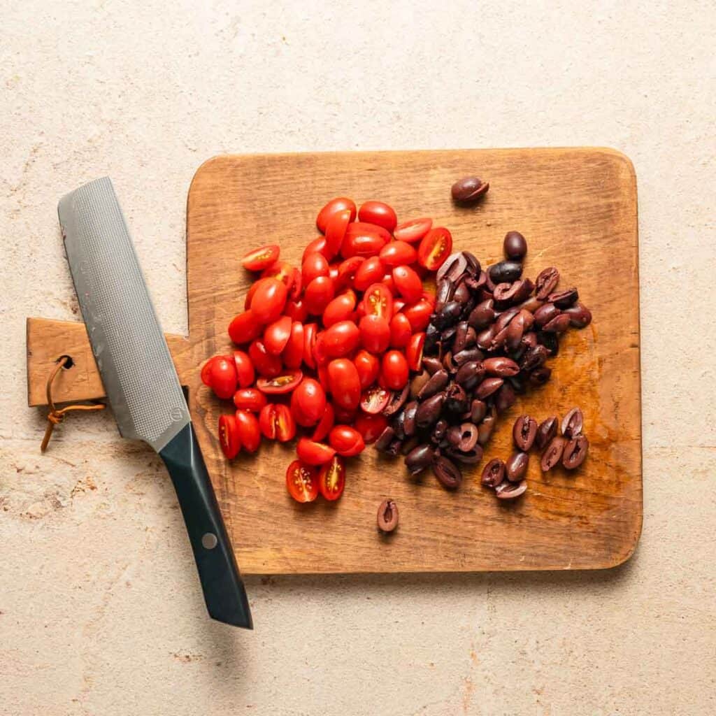 sliced olives and grape tomatoes on wooden cutting board with a knife on the side.