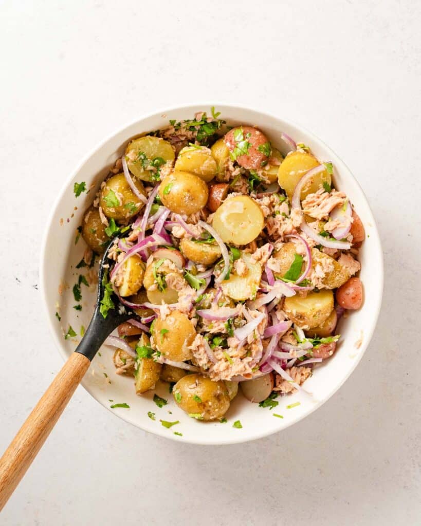 tuna and potato salad mixed in white bowl with wooden spoon.