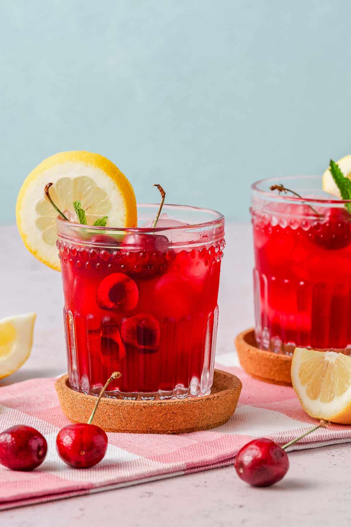 cherry lemonade in two glasses with lemon wedges and cherries on the side.