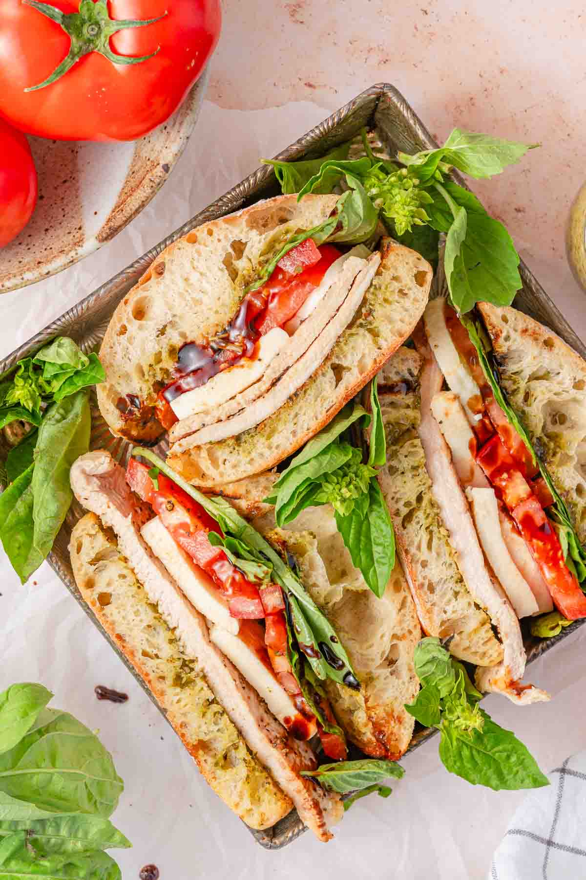 chicken caprese sandwiches in a metal tray with basil and tomatoes on the side.