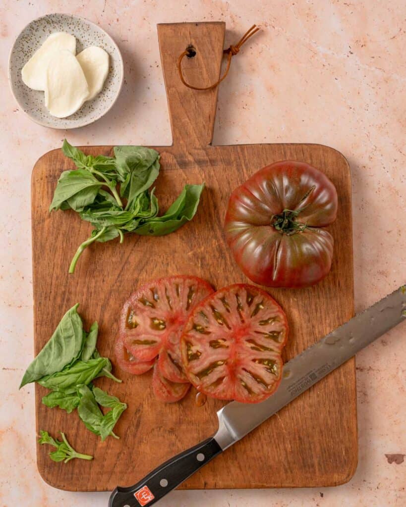 sliced tomato and basil leaves on cutting board with dish of fresh mozzarella cheese slices on the side.