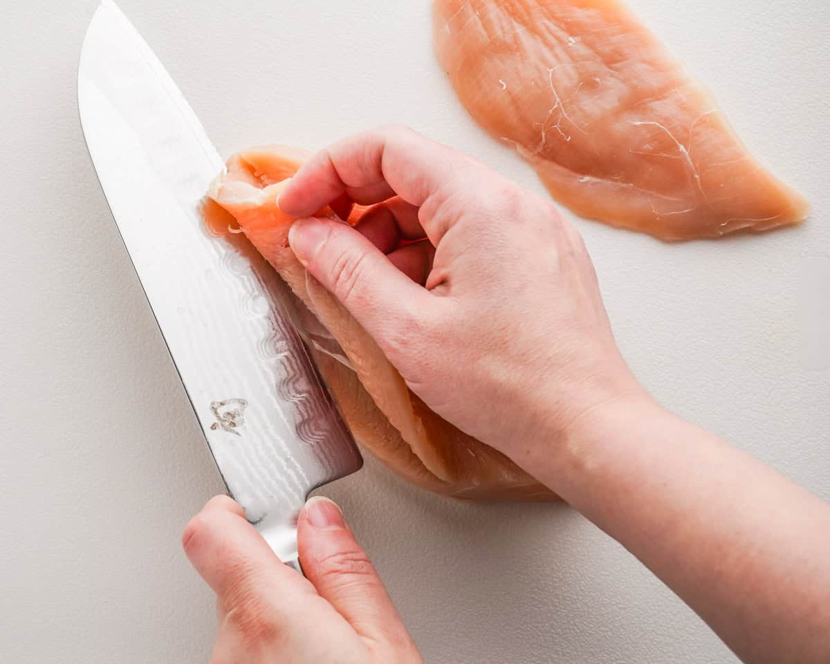 horizontal slice being cut from chicken breast with large knife.