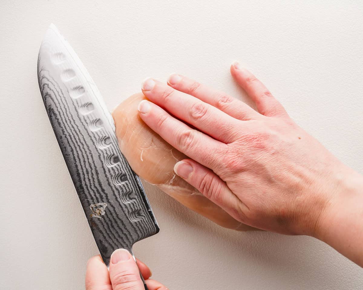 chicken breast being held in hand with knife beginning to slice it horizontally.