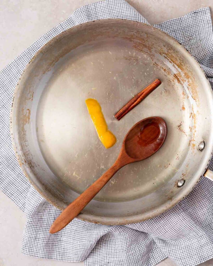 water sugar cinnamon stick and orange zest in pan with wooden spoon.