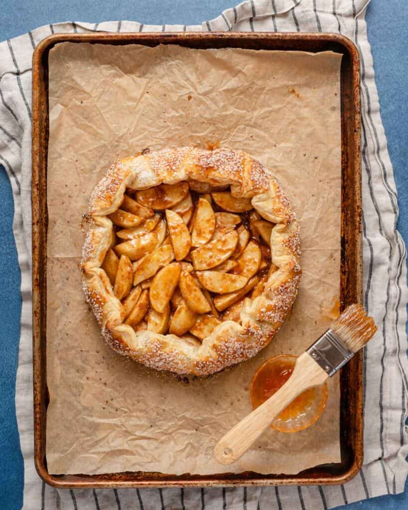 baked apple galette on baking tray with dish of apricot preserves and pastry brush on the side.