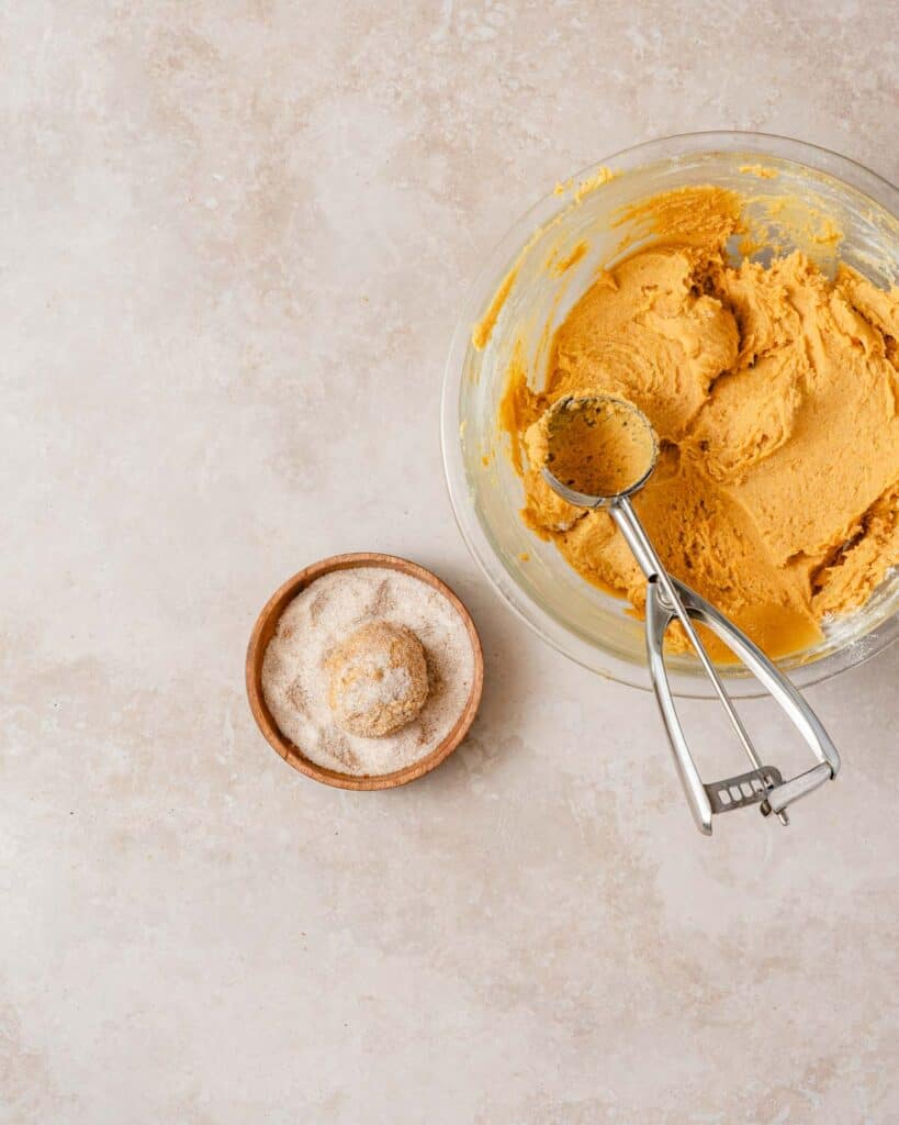 pumpkin cookie dough ball in dish of sugar with bowl of cookie dough and cookie scoop on the side.